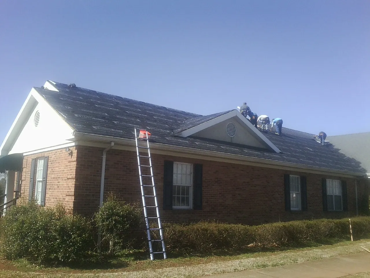 Crew tearing shingles off a brick ranch roof with a ladder set — Emergency Roof Repair work by our crew in Lamoni