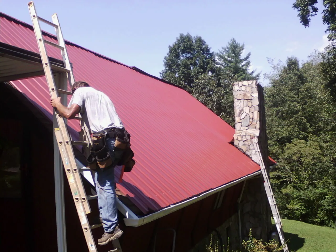 Roofer on a ladder beside a red metal roof and stone chimney — Soffit & Fascia Repair by Roofing Brothers in Lamoni